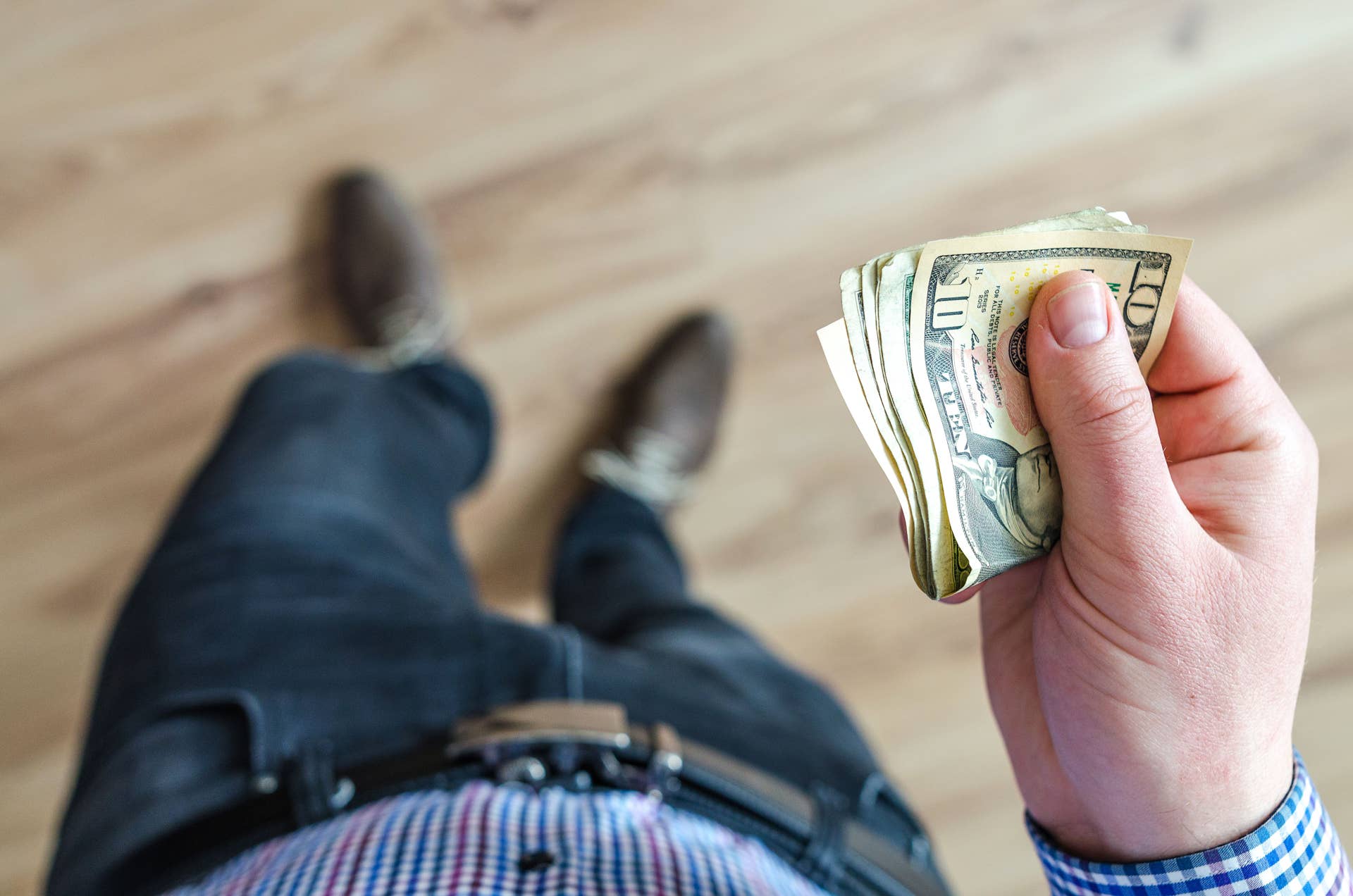 Close-up shot of a man holding US dollar bills indoors with a wooden floor background, Photo Credit: Lukas Blazek/Pexels