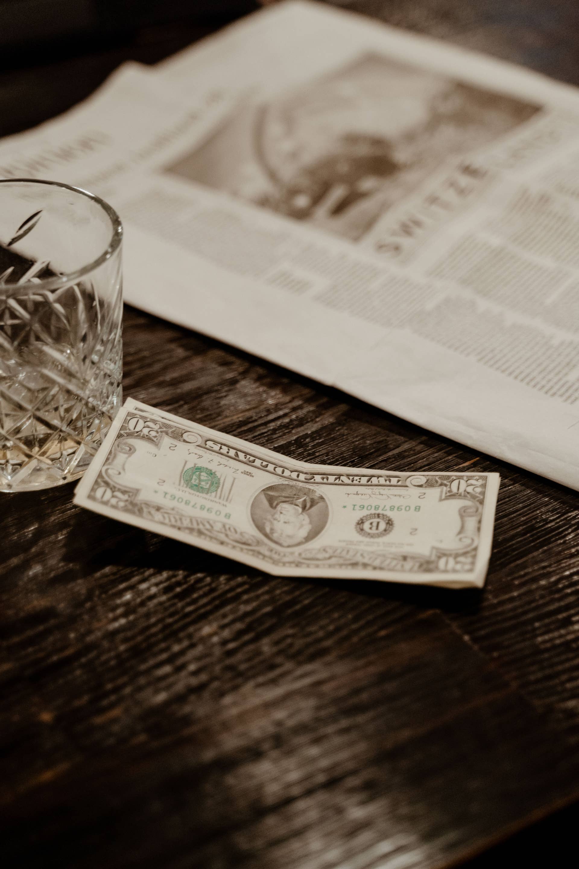 A vintage dollar bill next to a crystal glass on a wooden table and newspaper, Photo Credit: Mart Production/Pexels