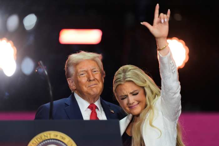 President Donald Trump standing with Erika Kirk who makes a hand gesture during Charlie Kirk's memorial, Photo Credit: Spitfire/X