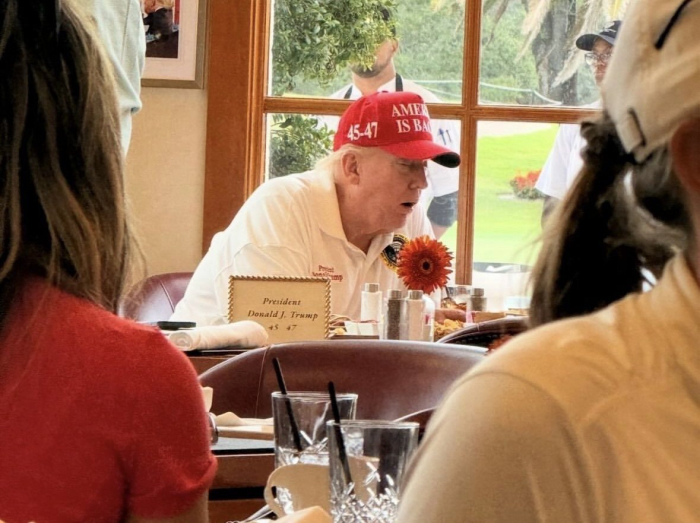 President Donald Trump sitting on a table at Mar-a-Lago wearing a red MAGA hat, Photo Credit: Socialist Planning Beyond Capitalism/X