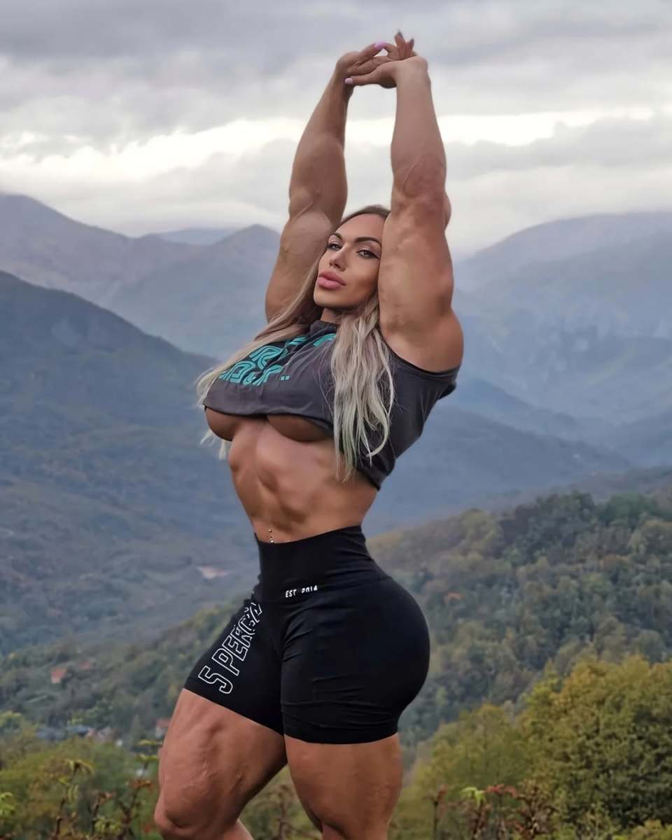 Natalia Kuznetsova flexing her arms above her head with a mountain landscape behind her, Photo Credit: nataliya.amazonka/Instagram