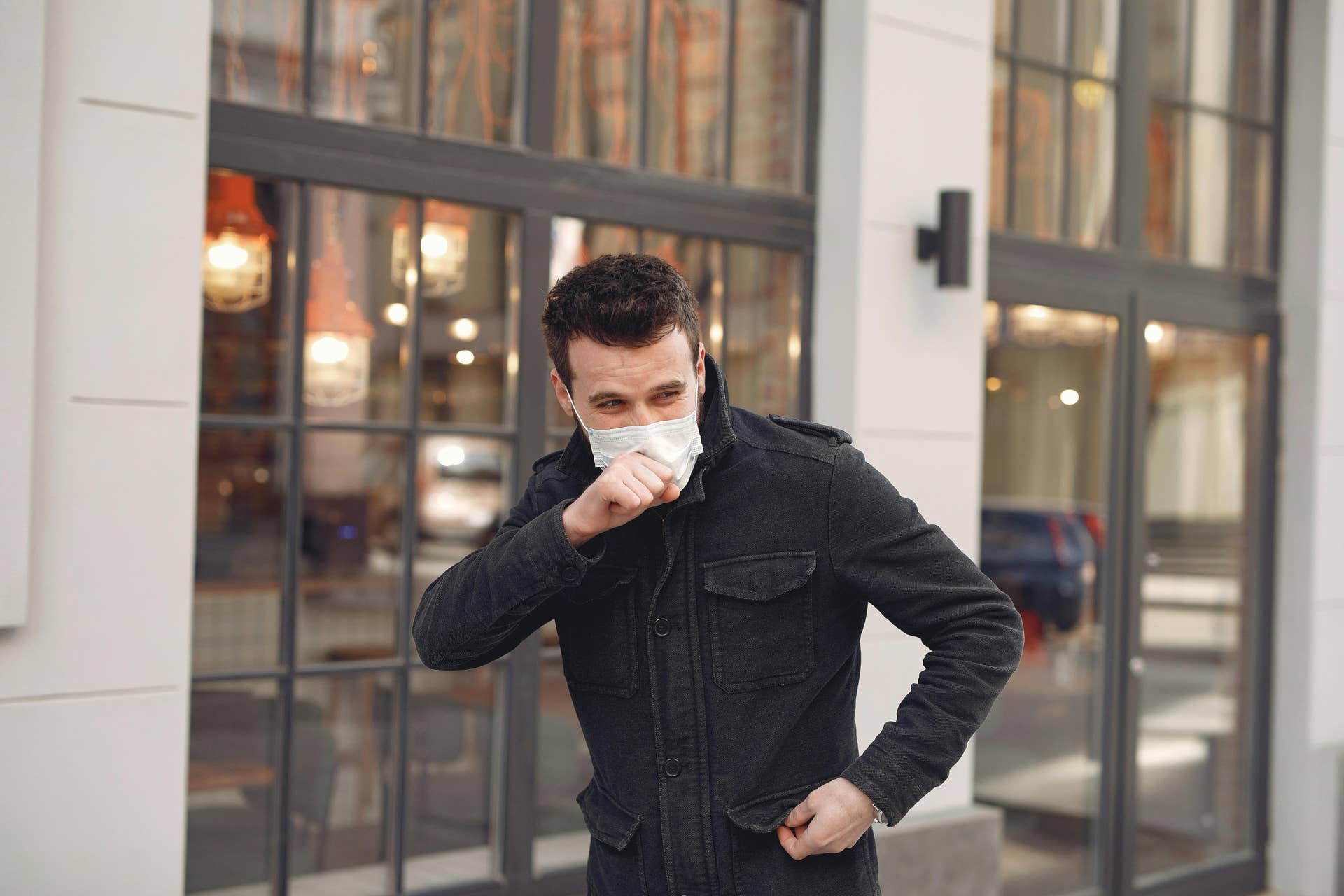 A man in a black jacket wearing a face mask while coughing, Photo Credit: Pexels